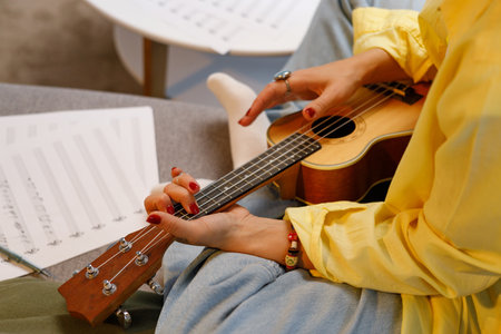 A beautiful hipster girl in a yellow shirt plays and sings on the ukulele on the sofa in the apartment. Composes a song, writes notes. Home Hobbies playing musical instruments.の写真素材