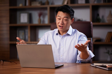 Asian man holding online webinar using laptop webcam, sitting in a home office. Man having online meeting and communicating remotely, looking at computer screenの写真素材