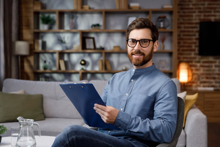 An adult handsome man sits at home in a stylish office. Psychologist, coach or businessman looking at the camera and smiling. Beautiful teeth and smile.の写真素材