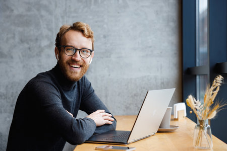 A young red-haired guy, a programmer or an entrepreneur in glasses, in a stylish cafe behind a laptop, looks at the camera and smiles. Freelancer works remotely. Online communication. small business.の写真素材