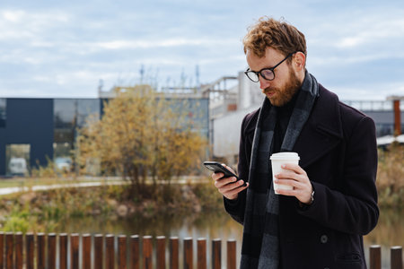 A young red-haired guy, a businessman in glasses, against the backdrop of urban style, checks mail on a smartphone, communicates in a messenger and drinks coffee. Online communication.の写真素材