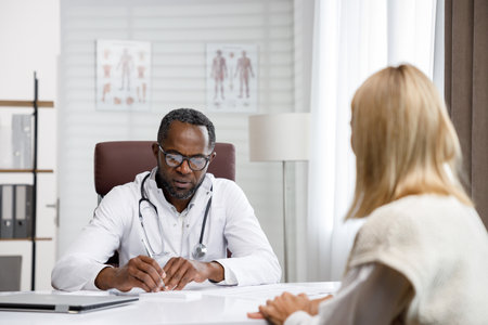 African American doctor, receives a patient, listens to complaints, makes diagnostics. Writes data. Doctors appointment.の写真素材