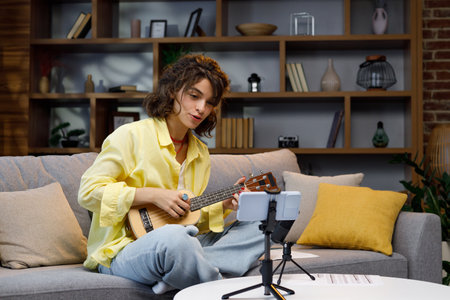 Blogger with mobile phone and microphone. A beautiful hipster girl in a yellow shirt plays and sings on the ukulele on the sofa in the apartment. Online learning to play the guitar.の写真素材