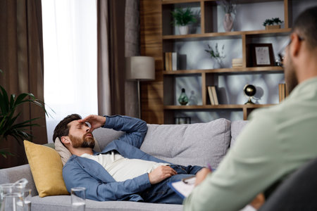 Visit to a psychologist. A handsome man is lying on the couch and talking to a psychotherapist. The patient is in depression, apathy and stress from burnout at work. Help of a psychologist.の写真素材