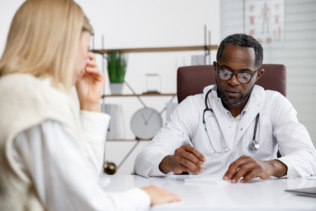 African American doctor, receives a patient, listens to complaints, makes diagnostics. Writes data. Doctors appointment.の写真素材