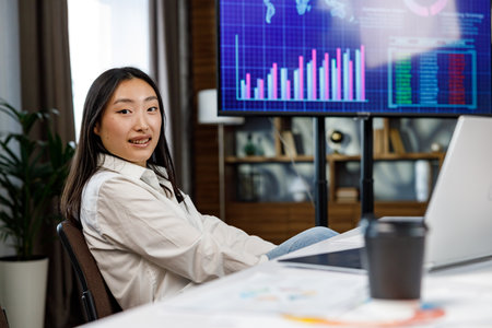 Confident happy smiling diverse asian woman, startup team leader. He sits at his workplace with a laptop, coffee on the background of a TV with charts and an analytical summary of marketing strategyの写真素材