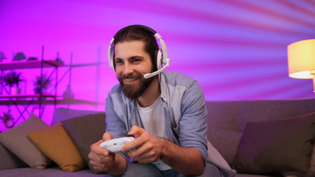 Cheerful young guy in a headset plays video games on the console while sitting on the couch in front of the TV. Gamer having fun together at home. Room with neon light.の写真素材