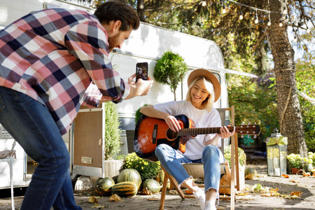 Funny and cheerful young couple. The girl plays the guitar, the guy photographs her for social networks against the backdrop of a travel trailer. Weekends in the summer time. Romance in nature.の写真素材