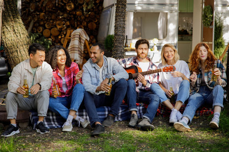 Handsome young man plays the guitar for friends. Mixed-race company of friends. Having fun listening to music at holiday summer parties near the travel trailer. Happy multi ethnic young people.の写真素材