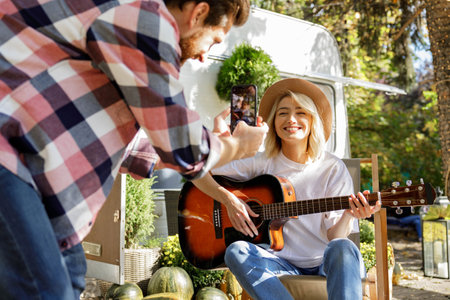 Funny and cheerful young couple. The girl plays the guitar, the guy photographs her for social networks against the backdrop of a travel trailer. Weekends in the summer time. Romance in nature.の写真素材