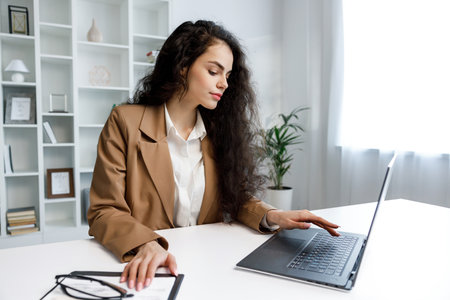 A young curly-haired brunette girl works at a laptop. Freelancer working from home, remote work. Successful manager girl, in a white office.の写真素材