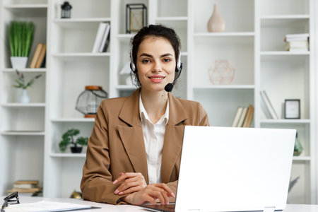 Young curly brunette woman wearing video call headset on laptop in home light office. Video conference, remote meeting. Programmer, remote work, work from home.の写真素材