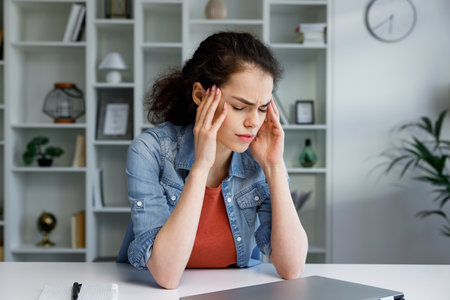 Young curly brunette woman sits stressed at the table, feeling apathy and depression from overwork at work. Burnout from the modern world. Problems in life, bad news. Headache, migraine.の写真素材