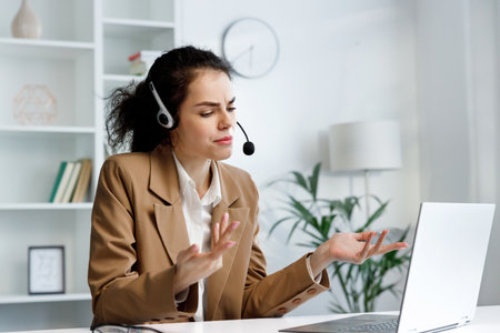Young curly brunette woman wearing video call headset on laptop in home light office. Video conference, remote meeting. Programmer, remote work, work from home.の写真素材
