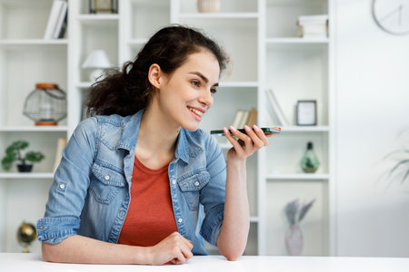 Young curly brunette sitting at the table uses a mobile phone. A voice message. Surfs the Internet, communicates in instant messengers on his smartphone. Online communication, smartphone in human lifeの写真素材