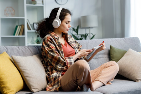 Curly-haired gir studentl sits on the sofa, wears headphones, looks in notebook and checks notifications on the phone. A beautiful woman studies and switches the music that will playの写真素材