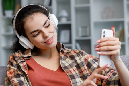 Young student girl in white headphones sitting on the sofa and taking a selfie in a beautiful bright apartment.の写真素材