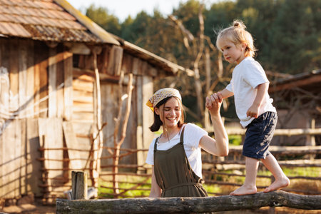Happy little kid with mother in domestic garden on warm sunny day. Parent supporting the child while he walking along the fence. Weekend with mom at the countryside on summer vacation. Love and careの写真素材