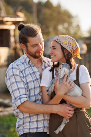 Portrait of young Caucasian happy farmer couple holding a little goat in their arms. Family business at ecological farm. Healthy food.Industrial production of goat milk dairy products. Cattle farming.の写真素材