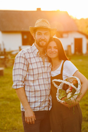 Portrait of young happy farmer couple holding basket with fresh organic vegetables on their rancho at the sunset. Vegetarian healthy food. Family business. Ecological farm and agriculture business.の写真素材