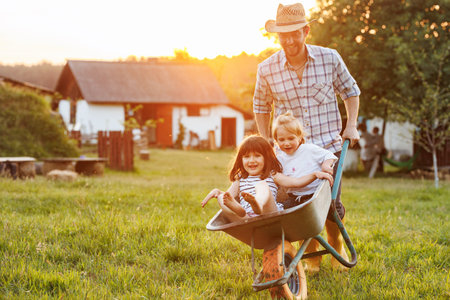 Happy little children having fun in a wheelbarrow pushing by dad in domestic garden on warm sunny day. Active outdoors games for family with kids. Spending time together on weekend at the countryside.の写真素材