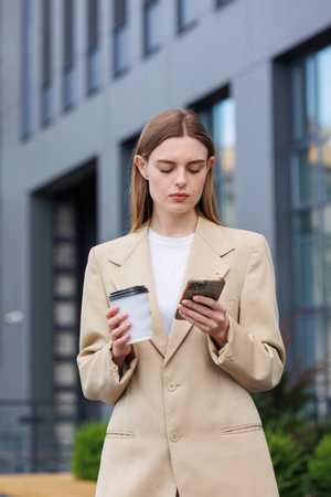 A young blonde girl against the backdrop of a business center, office center. Portrait of a successful startup. Smartphone, coffee casual suit.の写真素材
