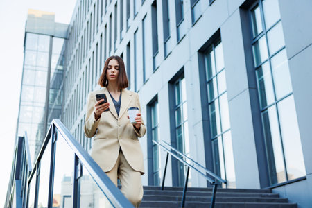 A young brunette girl against the backdrop of a business center, office center. On the go, he reads the news from his smartphone.の写真素材