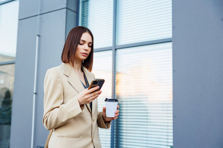 A young brunette girl against the backdrop of a business center, office center. Portrait of a successful startup. Smartphone, coffee casual suit.の写真素材