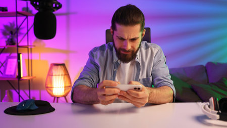 Gamer in neon room. A bearded guy sits at a table and plays a video game on his smartphone. Mobile video games. Rest and relaxation at home.の写真素材