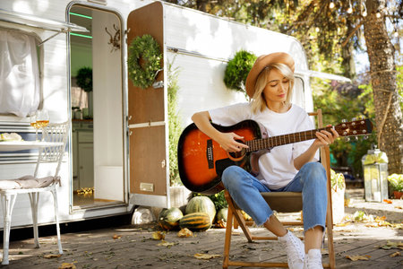 A young blonde girl with a guitar in front of a travel trailer. Rest outside the city. Happy people enjoy weekends in the summer.の写真素材