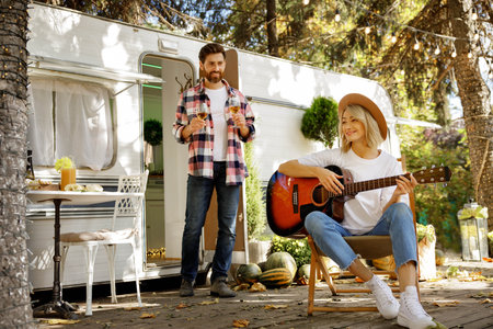 Lovely young couple on picnic near travel trailer. Blonde girl plays the guitar and guy brings out glasses of wine. Rest outside the city.の写真素材