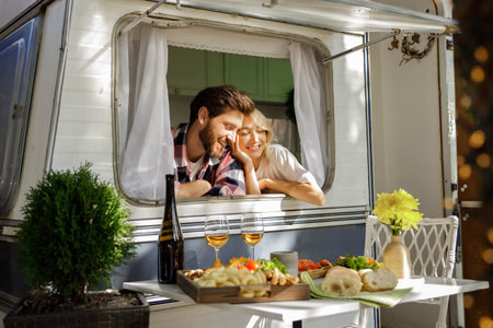 A lovely couple are standing in the sun, looking out the window of a tourist trailer, enjoying their vacation. People in love enjoying a sunny day outside the city, drinking wine.の写真素材
