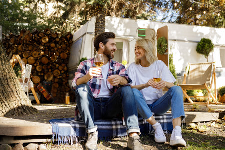 A lovely couple are sitting near the travel trailer, laughing having fun, enjoying their vacation. People in love enjoying a sunny day outside the city, drinking wine.の写真素材
