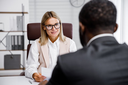 Hiring, interview. Middle-aged blond business woman conducting an interview in a bright office, an African American man. He looks at his resume and after reading it, shakes hands and smiles.の写真素材
