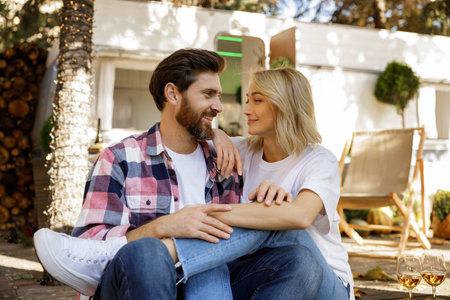 A lovely couple are sitting near the travel trailer, hugging, laughing having fun, enjoying their vacation. People in love enjoying a sunny day outside the city, drinking wine.の写真素材