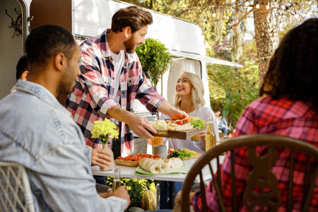 A cheerful multinational company is having fun on a picnic. Multisex friends sitting at the table outdoors celebrating eating and drinking drinks.の写真素材