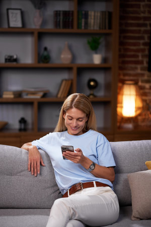 Blonde girl sitting on the couch looks into the smartphone, chatting, relaxing at home. Smiling adult woman communicates friendly by typing a message on the phone.の写真素材