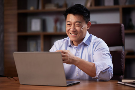 Asian man holding online webinar using laptop webcam, sitting in a home office. Man having online meeting and communicating remotely, looking at computer screenの写真素材