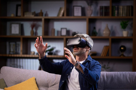 Adult grey-haired man using virtual reality and playing video games at home. Joyful man using virtual game and competing in virtual games while relaxing on sofaの写真素材
