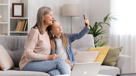 Grandmother senior lady and blonde little granddaughter taking selfie on phone sitting on sofa, looking at photos hugging.の写真素材