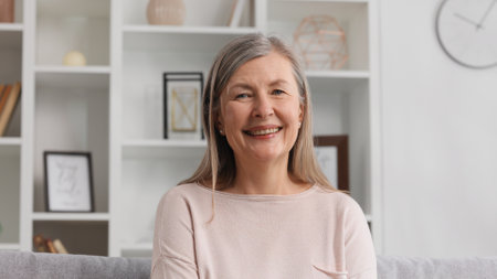 Happy senior woman sitting on sofa in nice apartment. A grayhaired grandmother in her 50s or 60s is resting on the couch at home, smiling.の写真素材