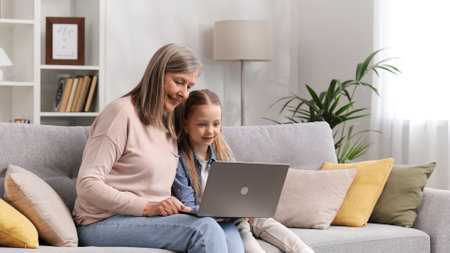 Grayhaired grandmother and little granddaughter are sitting on the sofa in the living room, resting and watching funny video cartoons on the laptop, talking, having fun.の写真素材