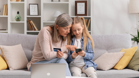 Young granddaughter teaches her senior grandmother to use a card for shopping on the Internet. A 60s grandmother and a little blond granddaughter are buying something by phone payment.の写真素材