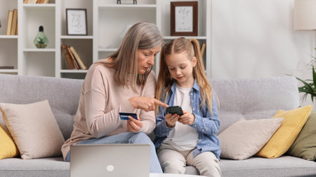 Young granddaughter teaches her senior grandmother to use a card for shopping on the Internet. A 60s grandmother and a little blond granddaughter are buying something by phone payment.の写真素材