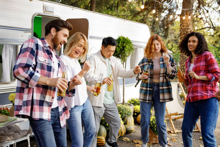 A cheerful group of friends rest in nature near a tourist trailer. Mixed-gender and mixed-race company of friends toasting, dancing, drinking drinks, have fun.の写真素材