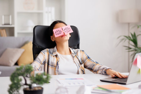 The concept of a lot of work. Tired Indian student girl sleeping with stickers on her eyes while sitting at the table. Distracted woman in home office.の写真素材