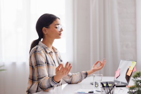 Managing stress in the workplace. Hindu student girl meditating and relaxing during a busy day while sitting at her workplace in an office interior. Focus on the hand.の写真素材
