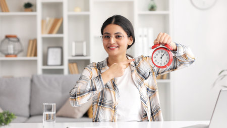 Indian smiling woman showing time on clock, approving, pointing finger at clock, encouraging. This is your time.の写真素材