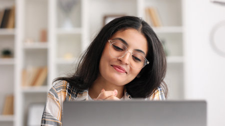 A young Indian girl works at a laptop and looks at the monitor. Beautiful woman talking on video call smiling. Studying in quarantine, listening to a podcast.の写真素材