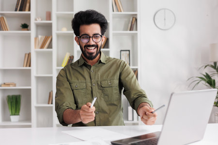 Satisfied Indian guy in black glasses checking his email and happy with good news. A young man winner dancing at the table celebrating the victory, waving pens in hands.の写真素材